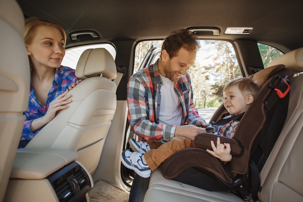 A father straps his son into his car seat while the mother watches from the front passenger seat.