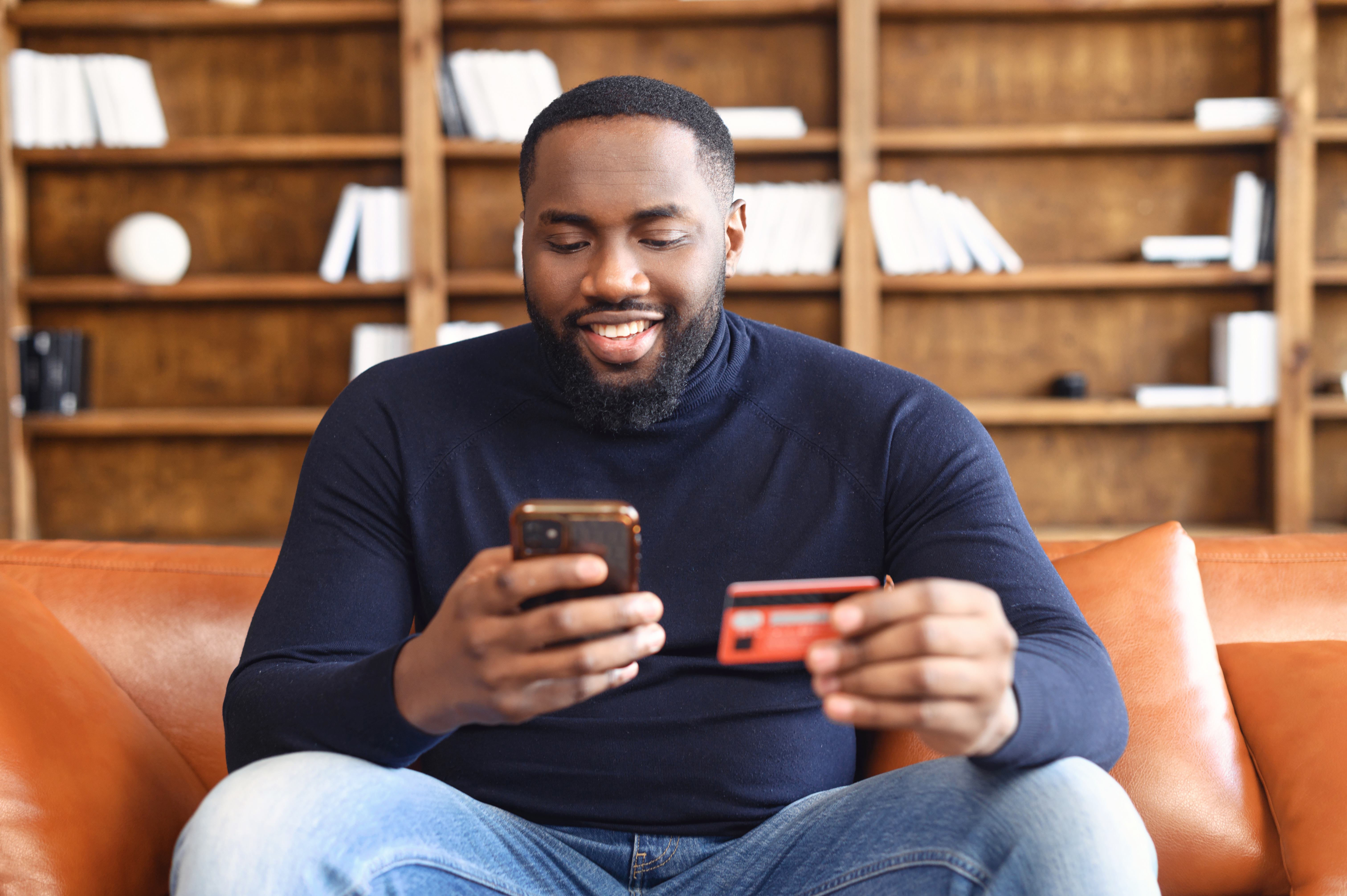 A man sits on his sofa and uses his credit card and mobile phone to manage his finances.